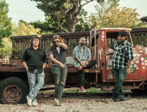 Four men lean against a rusted red pickup truck covered in stickers, posing outdoors in a yard.
