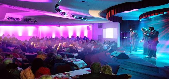 Crowd seated at round tables in a banquet hall watches a live band performing on a lit stage with purple and blue lighting.