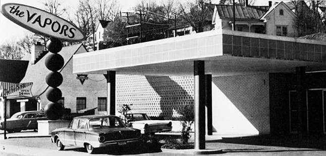 Black-and-white photo of a mid-century gas station with a large curved sign reading 'the VAPORS' and vintage cars parked near the pumps under a flat canopy.